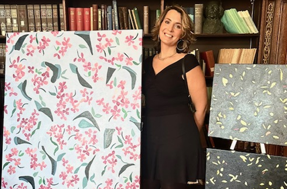 Ginger Mimmo Rohlfing stands next to a large painting of scattered green leaves and pink flowers, along with two smaller paintings with gray backgrounds and green leaves.